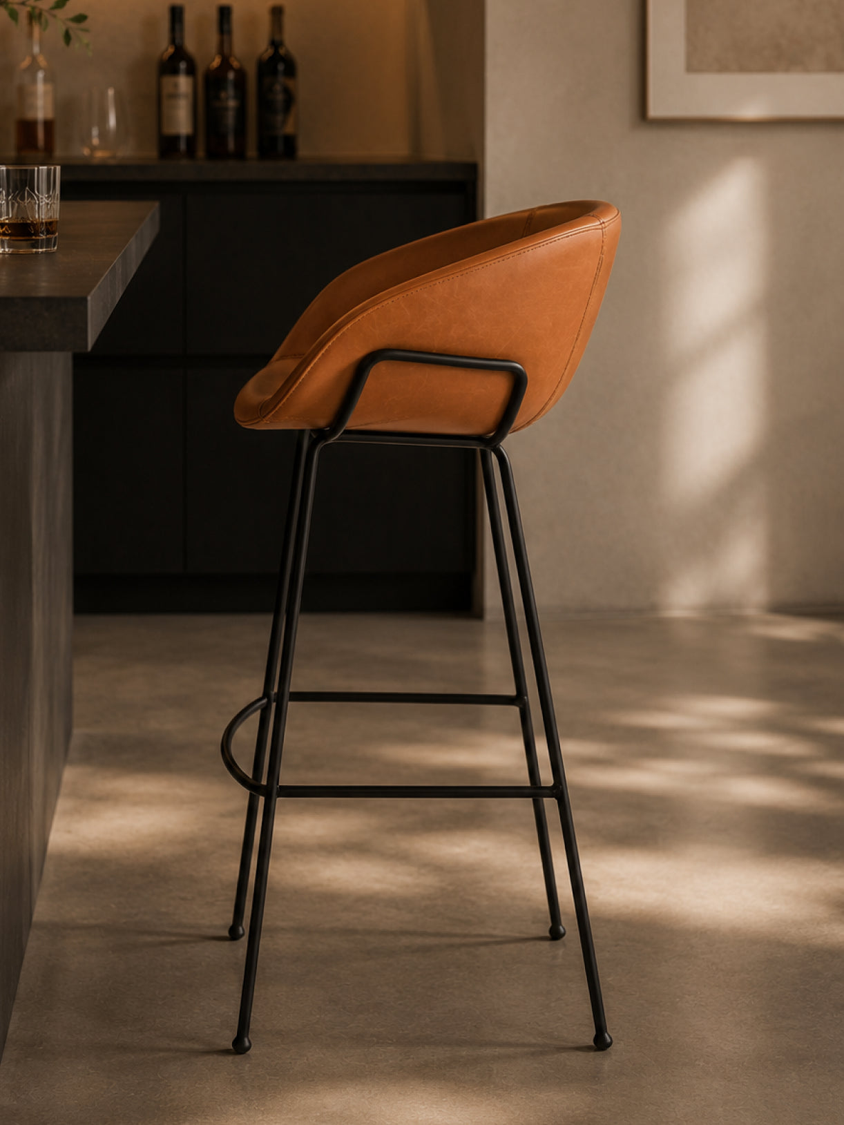 Barhocker Monkey in Seitenansicht mit cognacfarbenem Bezug, schwarzem Stahlgestell und minimalistischer Hausbar im Hintergrund